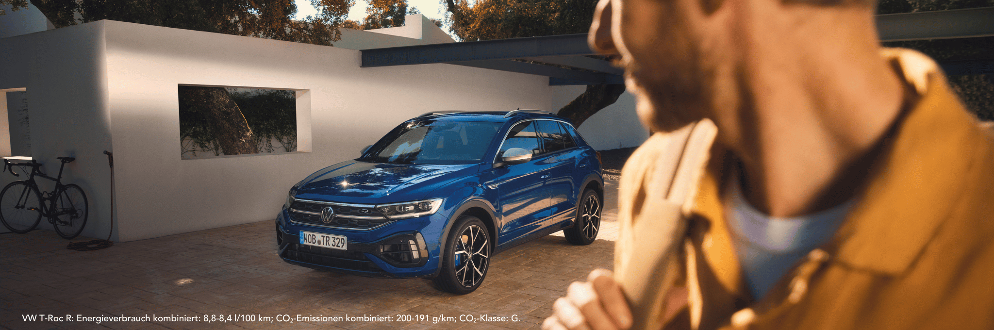 Blauer VW T-Roc R vor Haus und verschwomme Person mit Blick zum Fahrzeug