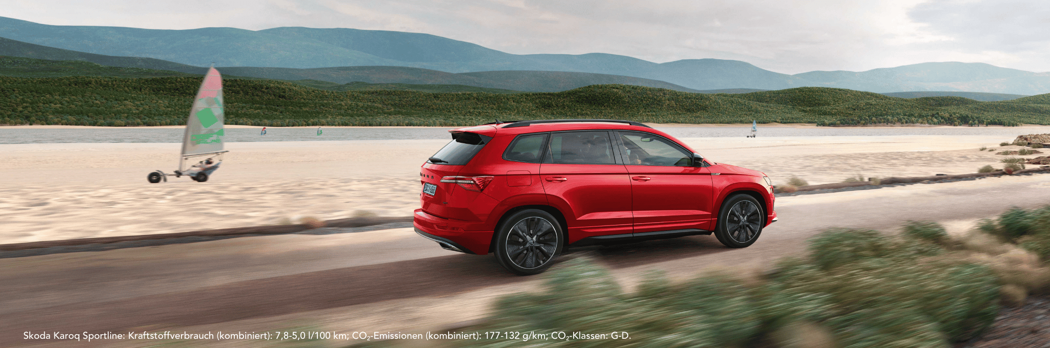 Ein roter Skoda Karoq Sportline fahrend vor einem Strand und etwas Wasser. Im Hintergrund sind Berge.